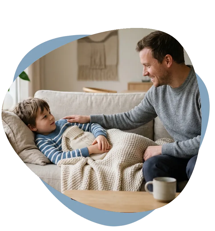 Vater tröstet seinen Sohn mit Bauchschmerzen, der mit Wärmflasche unter einer Decke auf dem Sofa liegt